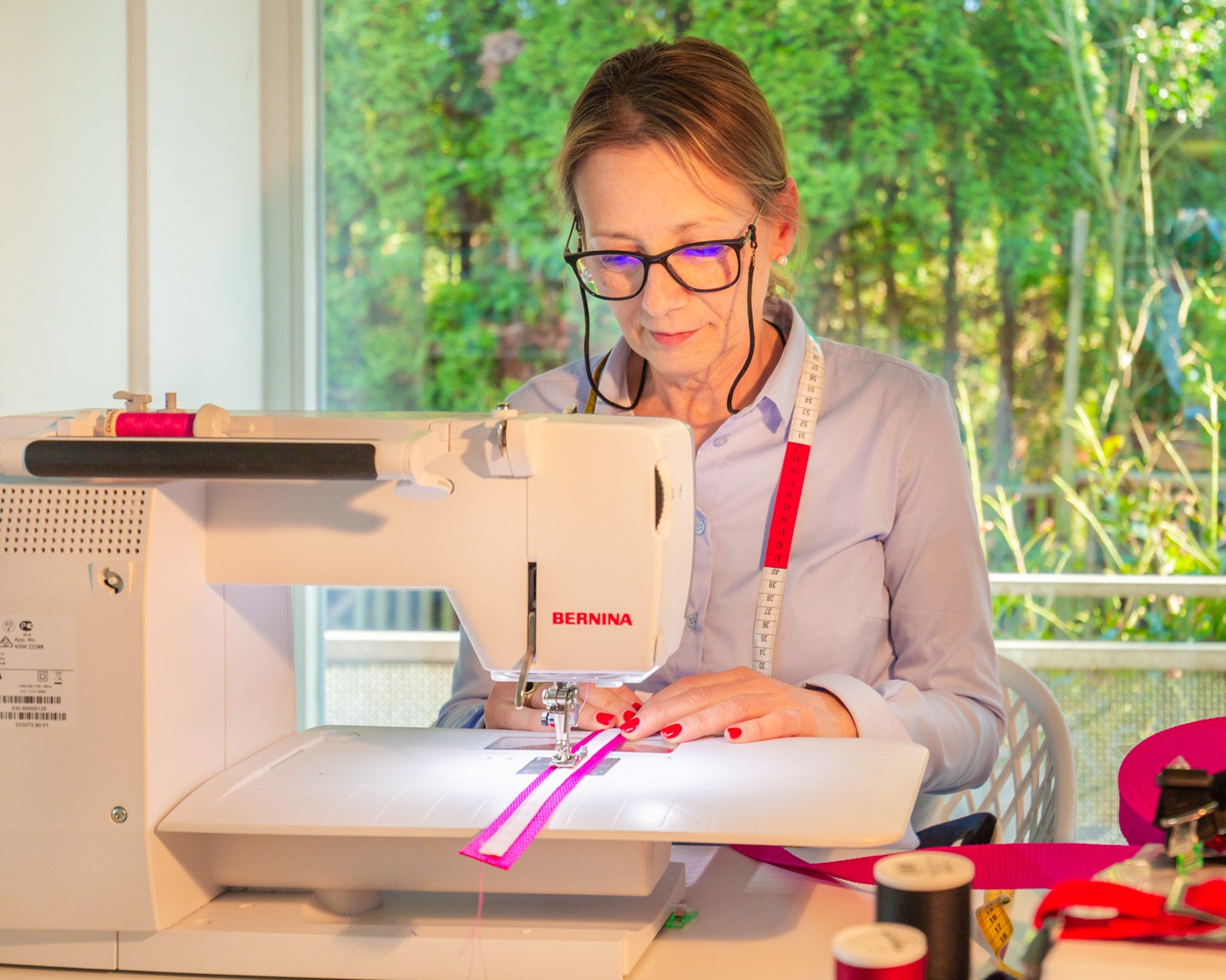 Ora Ebeling während der Arbeit sitzend an einer Nähmaschine mit Brille auf der Nase und Maßband über den Schultern.